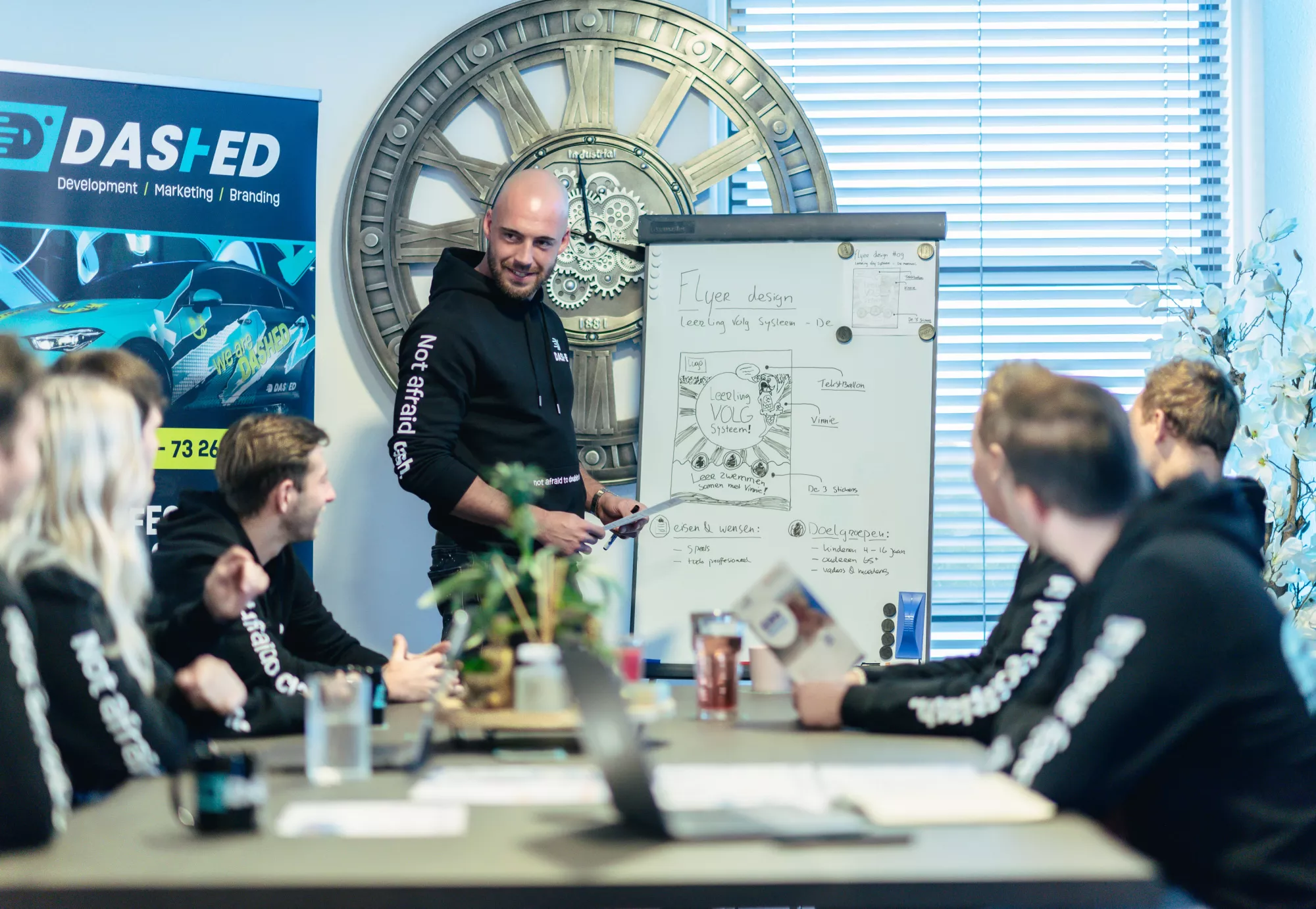 Een man presenteert ideeën aan een groep in een kantoor, staande bij een whiteboard met schetsen. Op de achtergrond een groot klokdecor en een poster van het bedrijf DASHED.