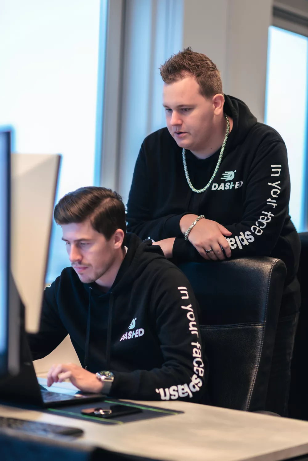 Twee mannen in zwarte hoodies overleggen achter een computerscherm in een kantooromgeving.