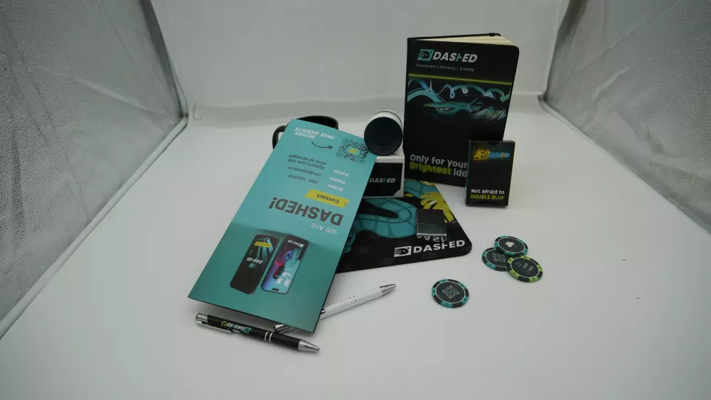 Promotiemateriaal op tafel met een flyer, pennen, een notitieboek, pokerchips en een mok, allemaal met het merk "DASHED".