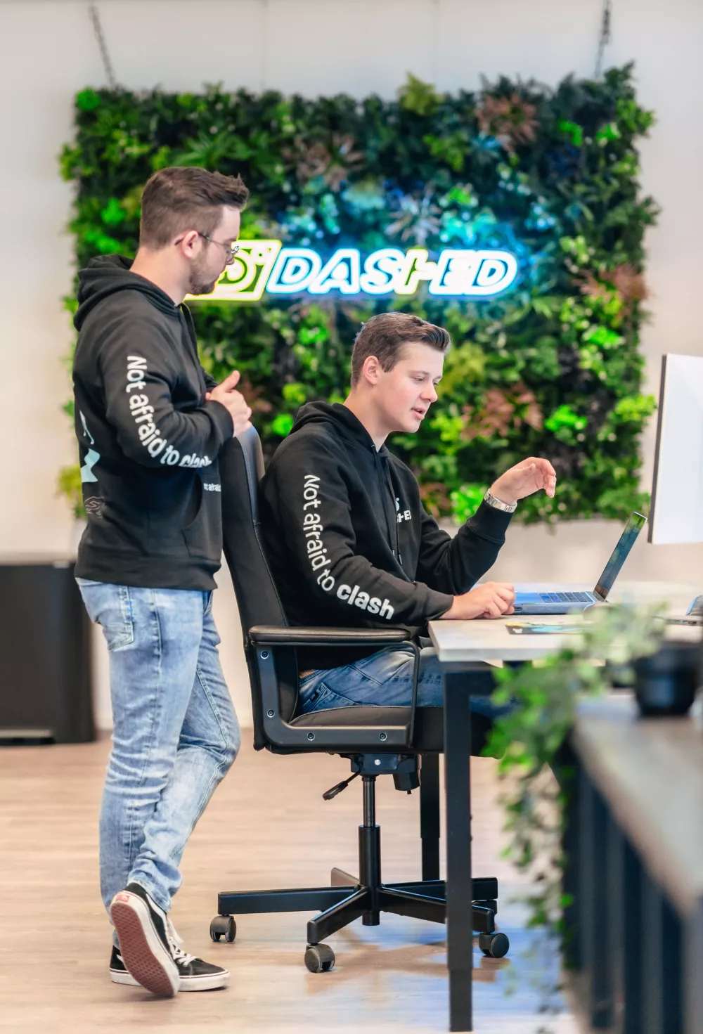 Twee personen in zwarte hoodies staan en zitten aan een bureau met een laptop. Op de achtergrond is een neonbord met de tekst "DASHED" voor een verticale plantenwand.