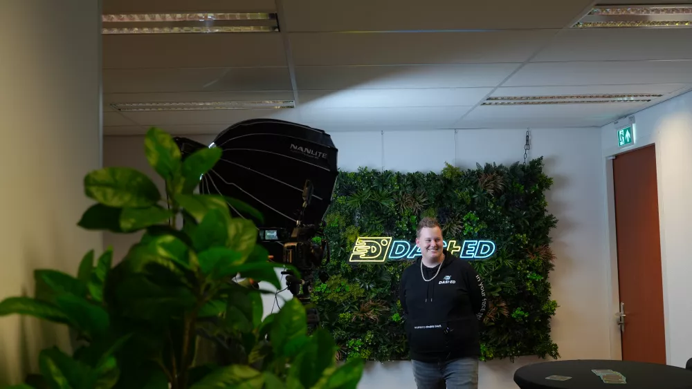 Een persoon poseert voor een met planten bedekte muur met een neonbord waarop "DASHED" staat, in een kantooromgeving met een opnamelicht en een camera op de voorgrond.