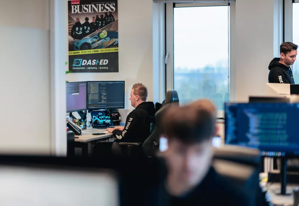 Kantooromgeving met programmatische code op beeldschermen. Medewerkers werken geconcentreerd achter computers. Poster aan de muur en zicht op een raam met uitzicht naar buiten.