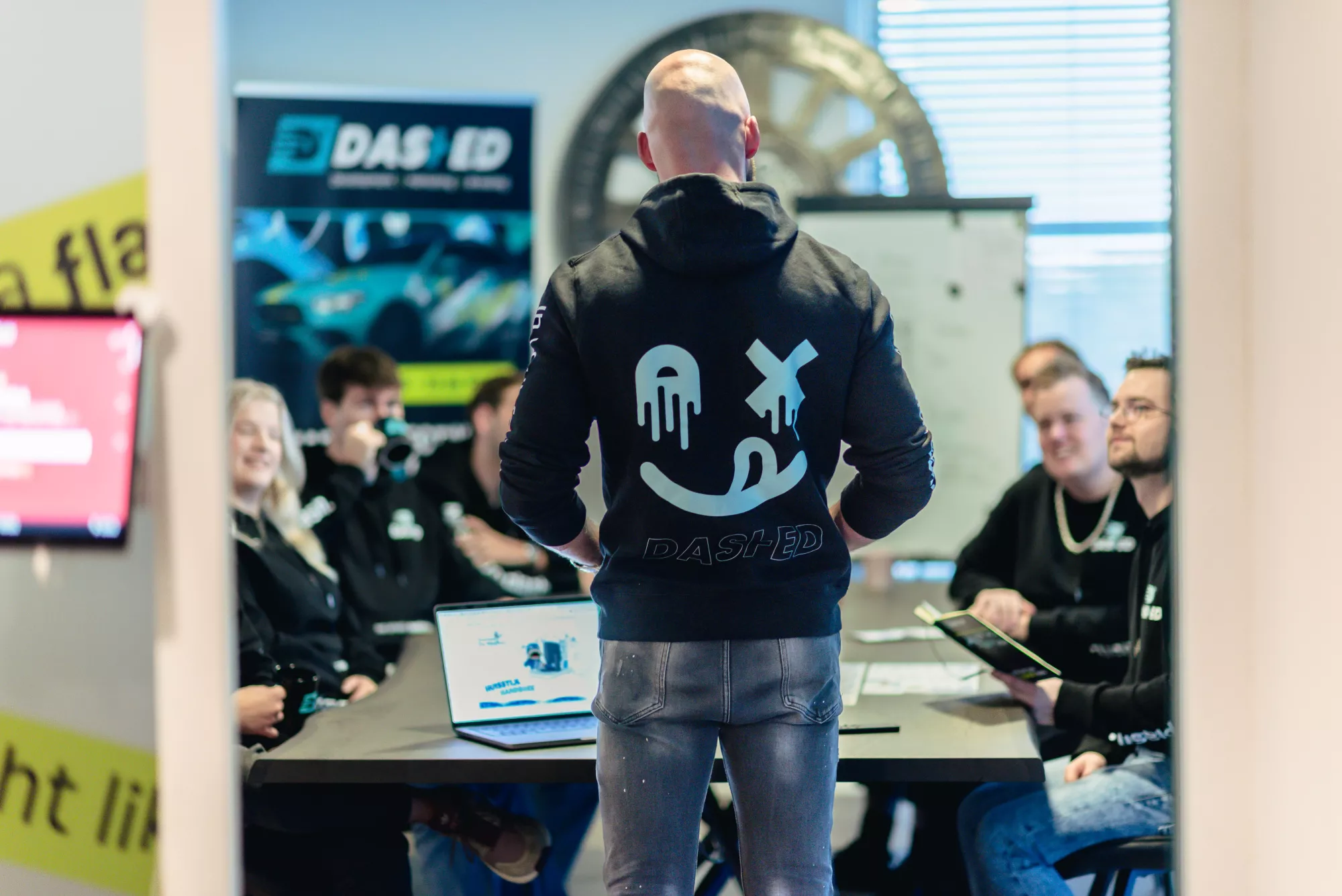 Een man staat met de rug naar de camera en spreekt een groep mensen toe die rond een tafel zitten met laptops en notities. Op zijn trui staan grafische symbolen en de tekst "DASHED".