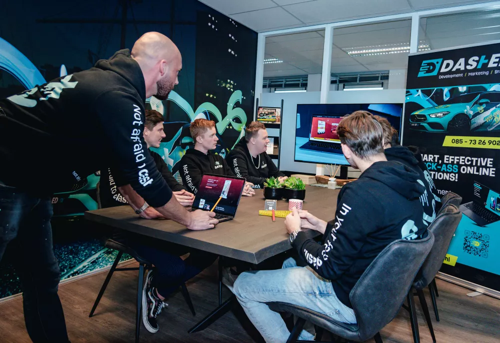 Een groep mensen in hoodies met teamlogo zit aan tafel en kijkt naar een presentatie op een groot scherm in een moderne vergaderruimte. Er zijn laptop en reclamemateriaal aanwezig.