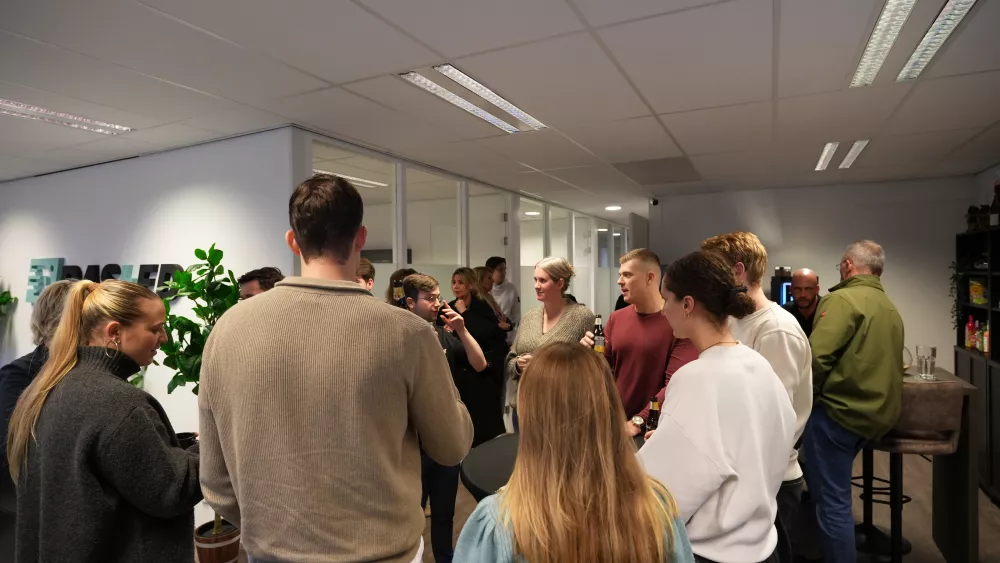Een groep mensen staat bij elkaar in een moderne kantoorruimte, pratend en drinkend, met enkele planten en glazen wanden op de achtergrond.