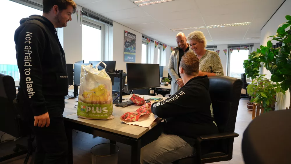 Mensen in een kantoor vieren samen een gelegenheid. Iemand zit voor een bureau met cadeauverpakking, terwijl anderen glimlachend toekijken. Een Plus-zak met bananen staat op tafel.