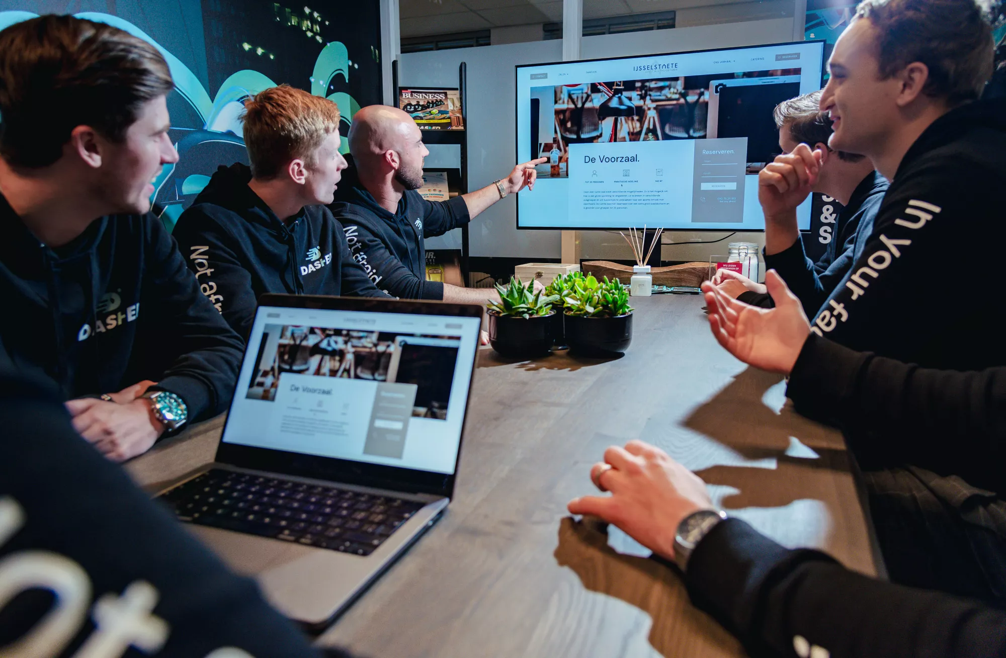 Een groep mannen zit rond een tafel tijdens een vergadering, waarbij een man naar een scherm wijst dat een website toont. Op de tafel staan laptops en planten.