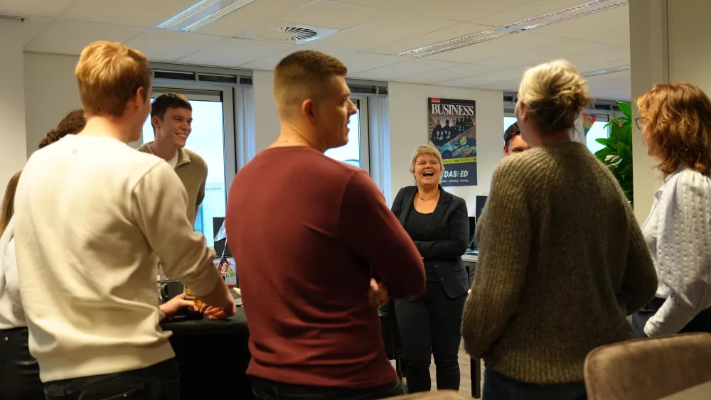 Een groep mensen staat in een kring in een kantoor, lachend en pratend met elkaar. Een poster met "Business" hangt aan de muur.