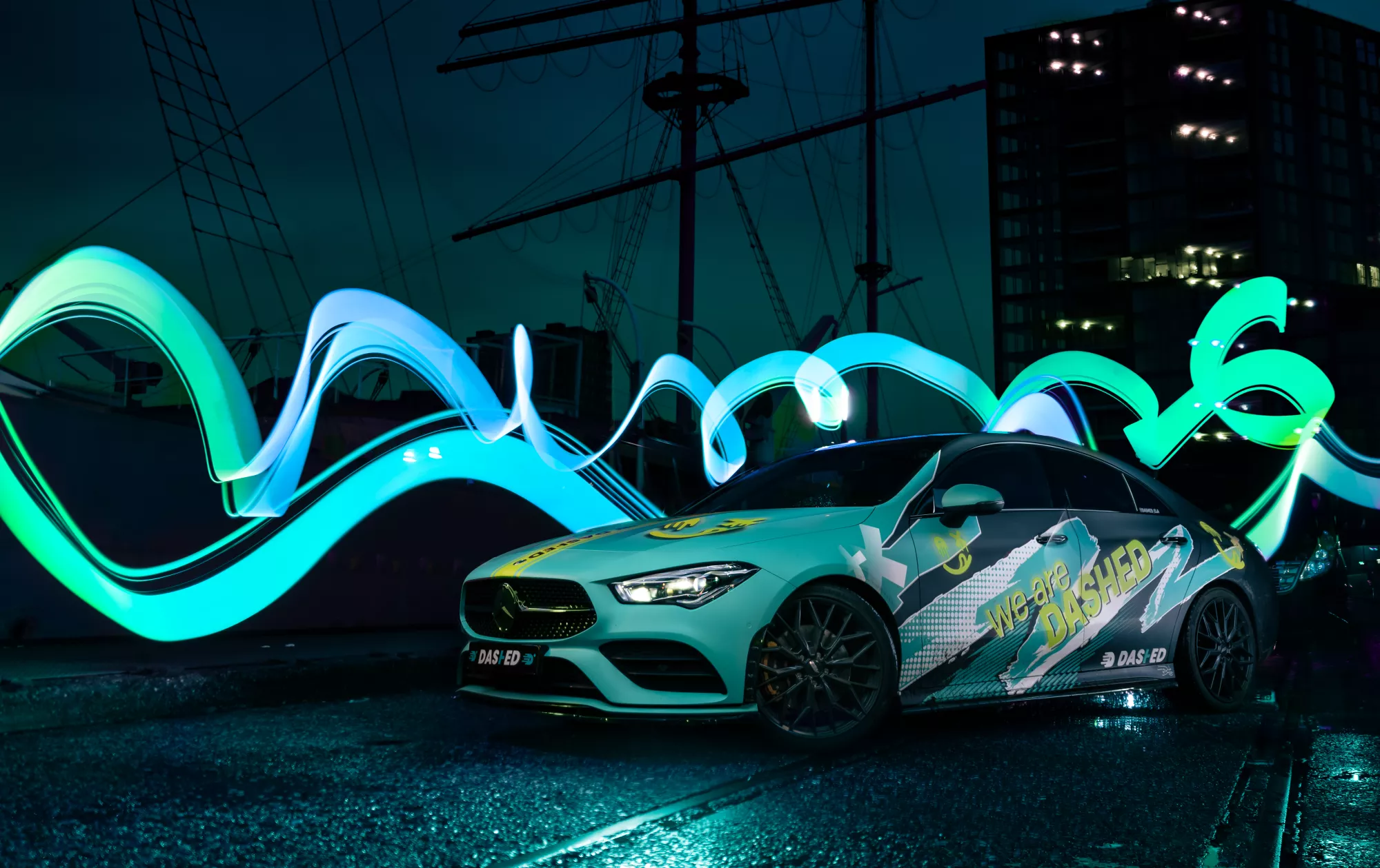 Witte auto met grafische stickers staat op een natte kade voor een schip en gebouw. Heldergroene en blauwe lichtstrepen dansen rondom, wat een futuristische sfeer creëert.
