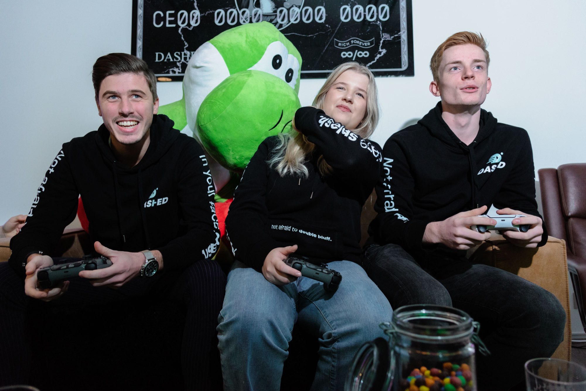 Drie collega's gamen samen op de bank met de groene mascotte ertussen
