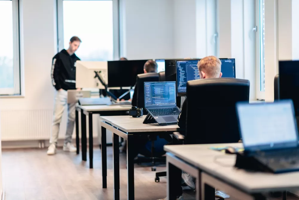 Een moderne kantooromgeving met meerdere personen die aan bureaus werken met computers. Enkele schermen tonen programmeercode.