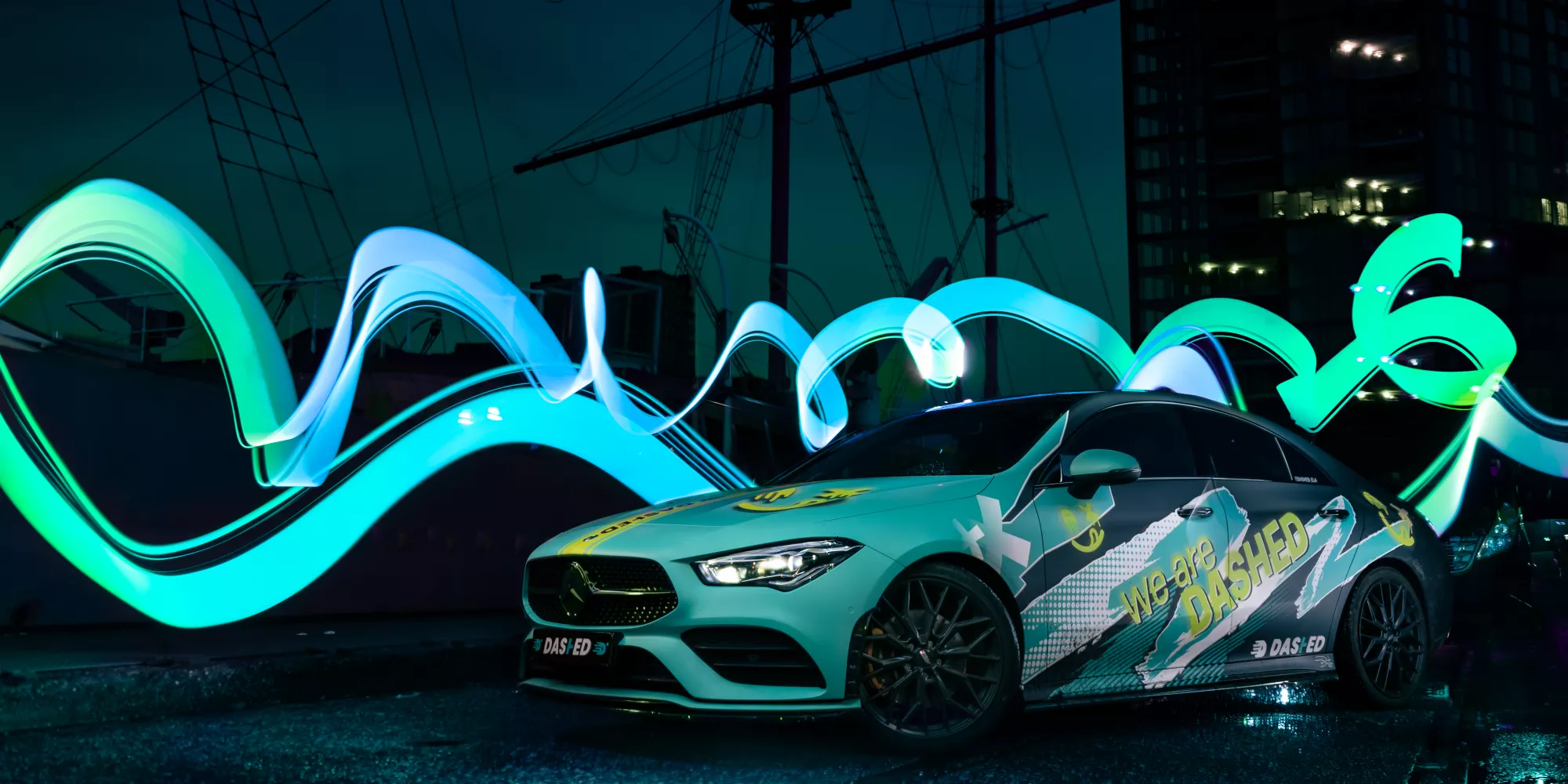 Een sportieve auto staat geparkeerd voor een nachtbewolkte lucht met een zeilschip en een gebouw op de achtergrond. Heldere blauwe en groene lichtstrepen dansen door de lucht rondom de auto, die gedecoreerd is met grafische ontwerpen en teksten.
