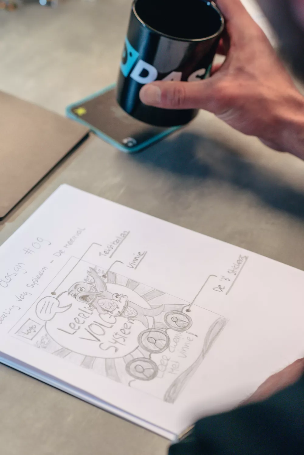 Hand schetst een ontwerp op papier met tekst en illustraties, naast een hand die een zwarte mok vasthoudt, op een bureau.