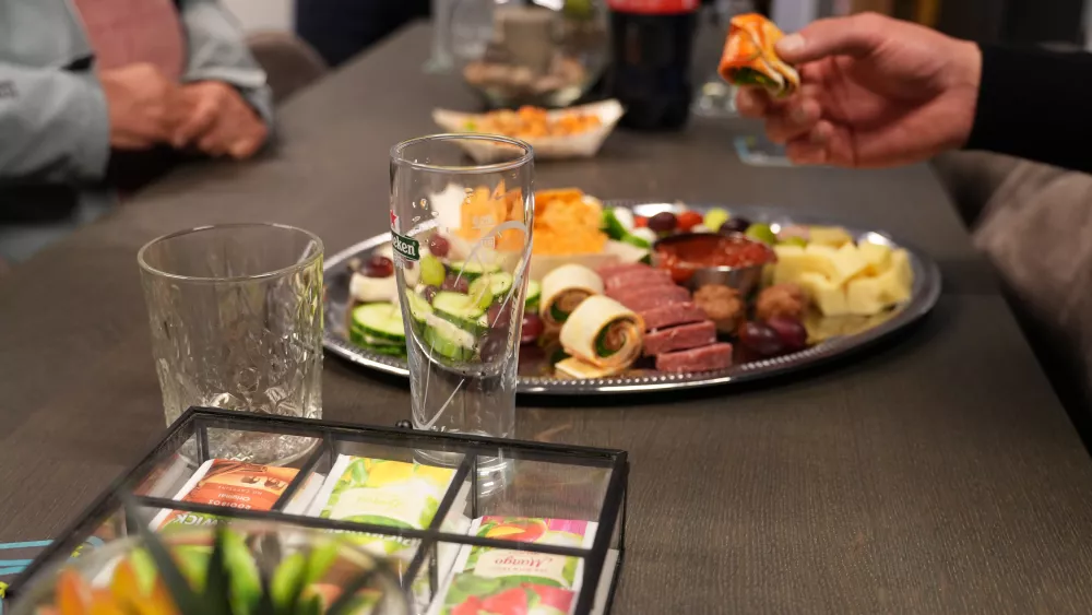 Een tafel met een schaal vol hapjes zoals kaas, vleeswaren en druiven. Een persoon houdt een stukje zalmrol vast. Op de voorgrond staan twee lege glazen en een theedoosje.