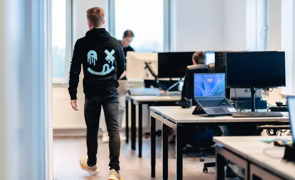Een persoon loopt in een moderne kantoorruimte met werkplekken, laptops en monitoren. Op de achtergrond werken mensen aan hun bureaus.