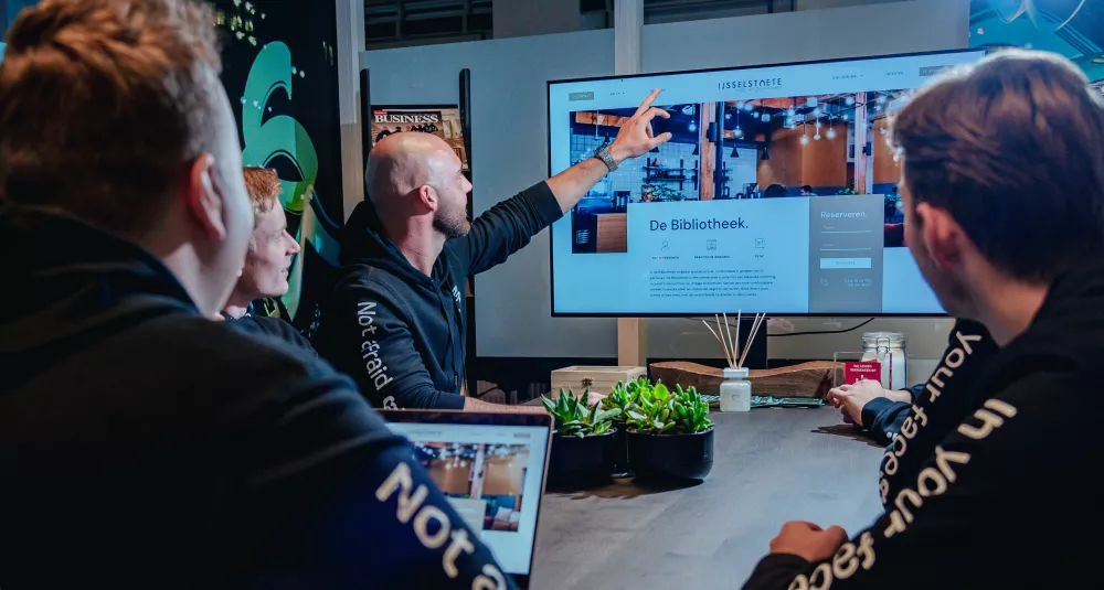 Mensen in een vergaderruimte kijken naar een scherm waarop een website wordt gepresenteerd. Een man wijst naar het scherm terwijl anderen aandachtig toekijken.