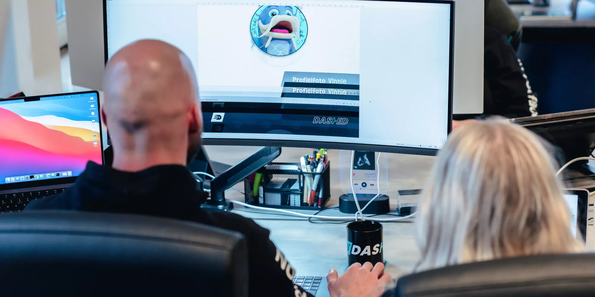 Twee mensen werken achter een bureau met grote computerschermen, bezig met een ontwerp van een vislogo. Op de achtergrond bevinden zich bureau-accessoires en laptops.