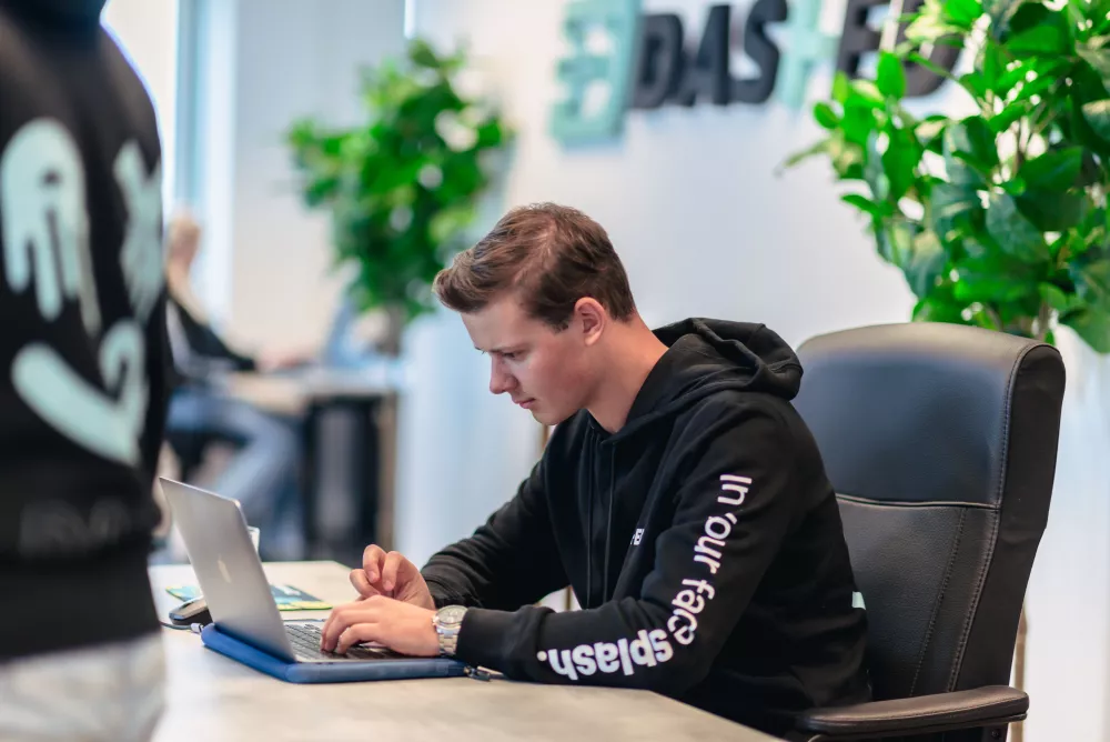 Man in zwarte hoodie werkt geconcentreerd op een laptop in een modern kantoor met planten.