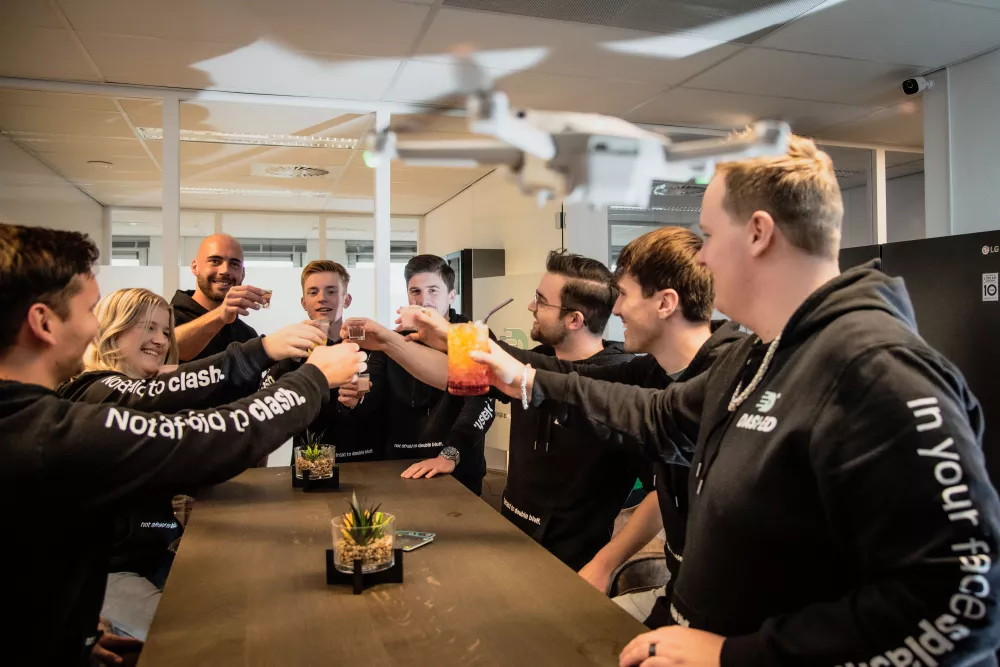 Een groep mensen in zwarte hoodies proost samen met drankjes in een modern kantoor. Op de voorgrond is een drone te zien.