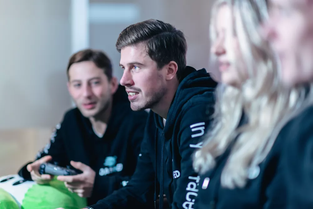 Drie mensen in zwarte hoodies zitten naast elkaar; de persoon links heeft een gamecontroller vast, terwijl de anderen aandachtig naar voren kijken.