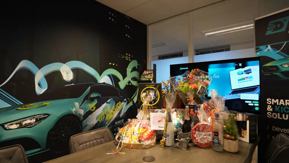 Bureau met cadeaumanden, bloemen en flessen in een kantoor met een muurprint van een auto en een groot scherm.