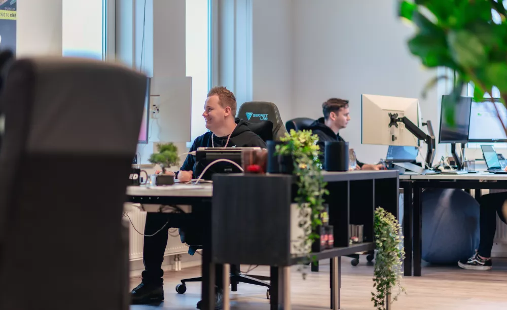Twee mensen werken aan bureaus met computers in een modern kantoor, omringd door kamerplanten.