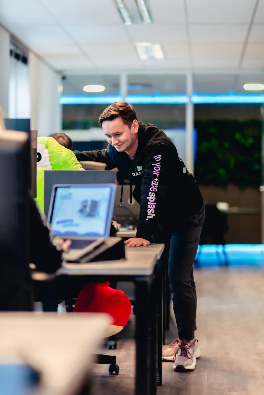 Een man leunt over een bureau in een kantoor en kijkt naar een computersysteem. Op de achtergrond is een groen knuffeldier te zien.