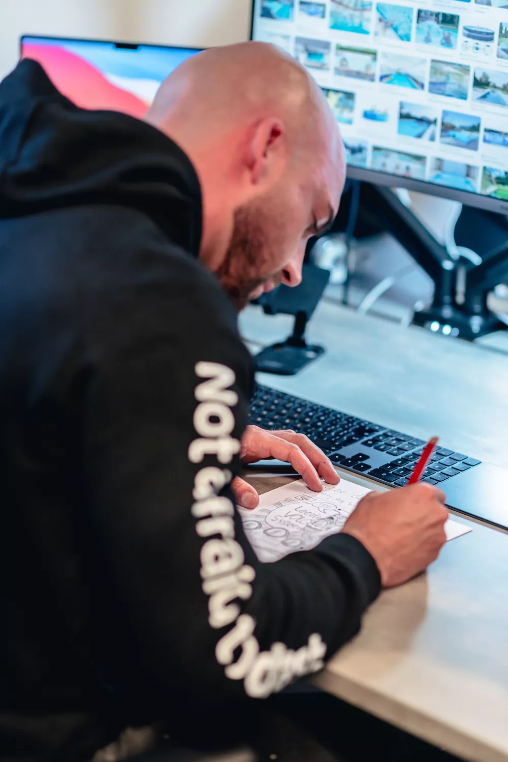 Een persoon tekent met potlood op papier aan een bureau met een computer waarop afbeeldingen te zien zijn.