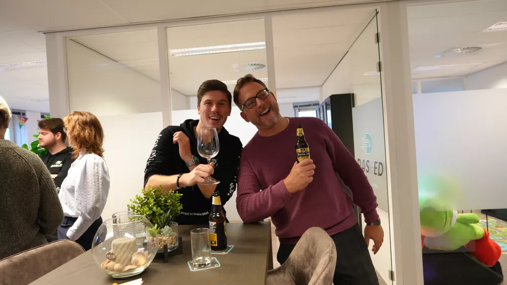 Twee lachende mannen poseren in een kantooromgeving met drankjes in de hand. Op de achtergrond staan enkele mensen en planten.