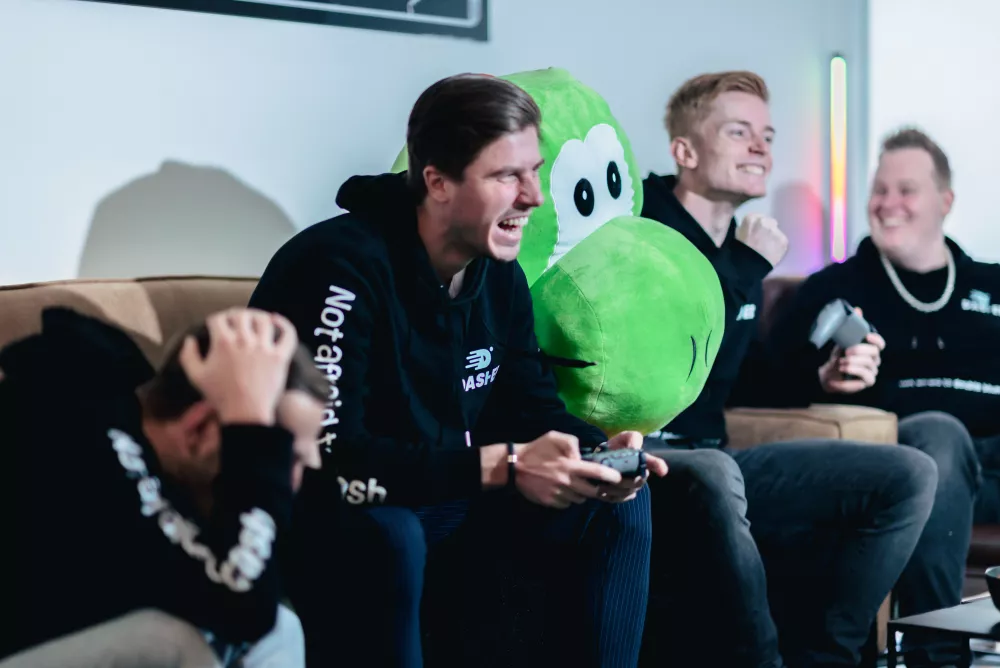 Vier mannen zitten op een bank en spelen videogames, waarvan er een een grote groene knuffel vasthoudt. De sfeer is vrolijk en competitief.