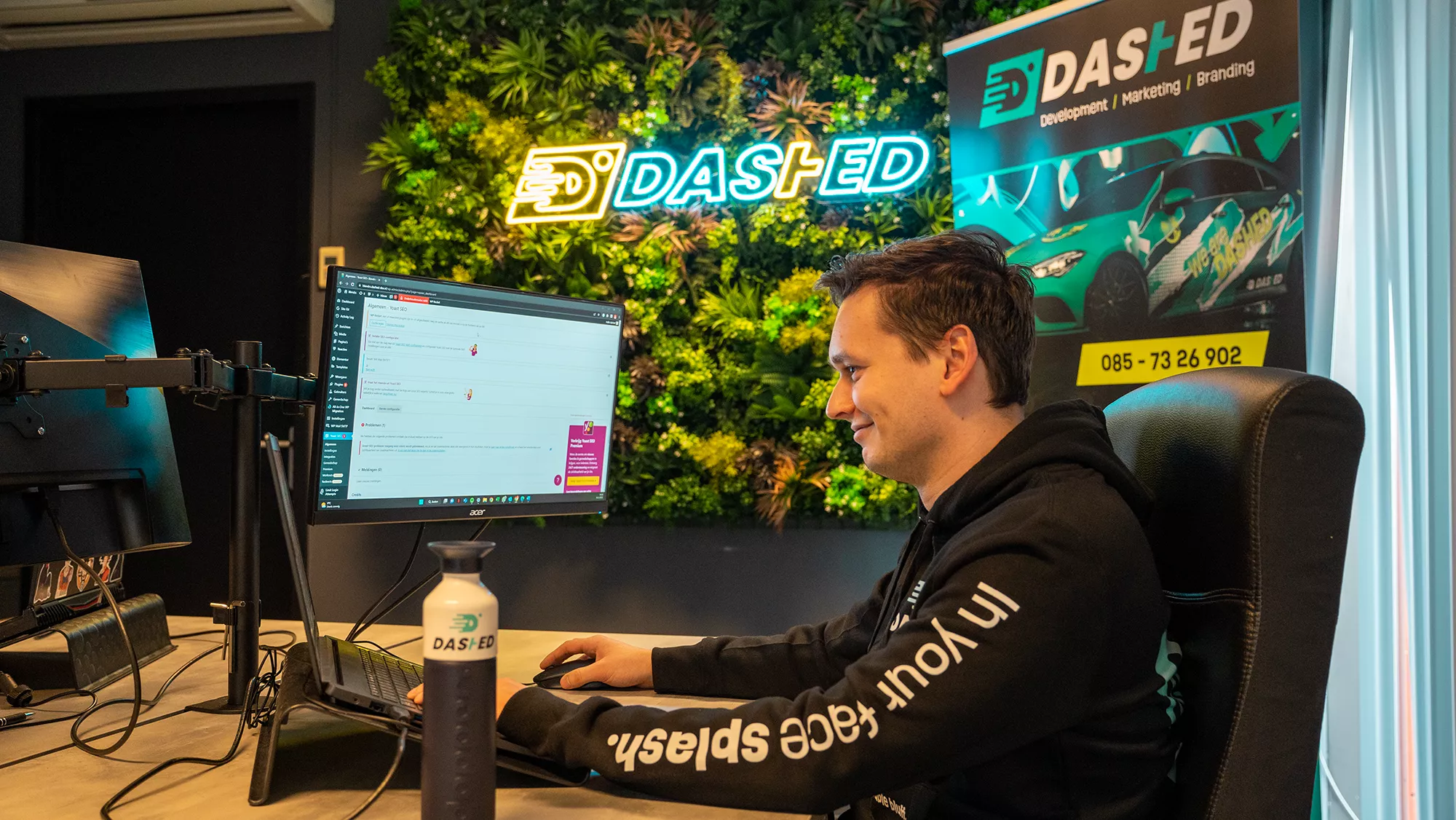 Man werkt aan een computer in een kantoor met een verlichte "DAS-ED" logo op de achtergrond en promotiemateriaal voor ontwikkeling, marketing en branding.