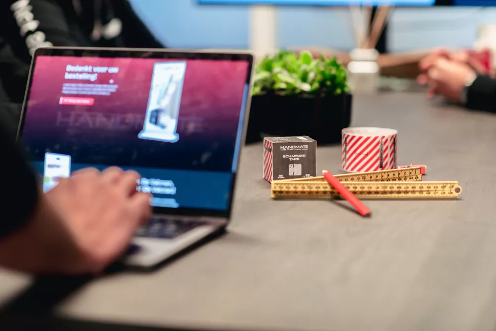 Een persoon werkt op een laptop aan een tafel met een rolmaat, een pen, en een doos Scharnier Tape naast een plant.