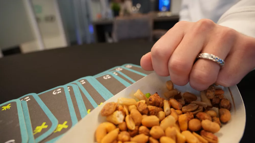Een hand met een ring pakt een snackmix in een bakje op een tafel, naast een stapel kaarten met blauwe randen.