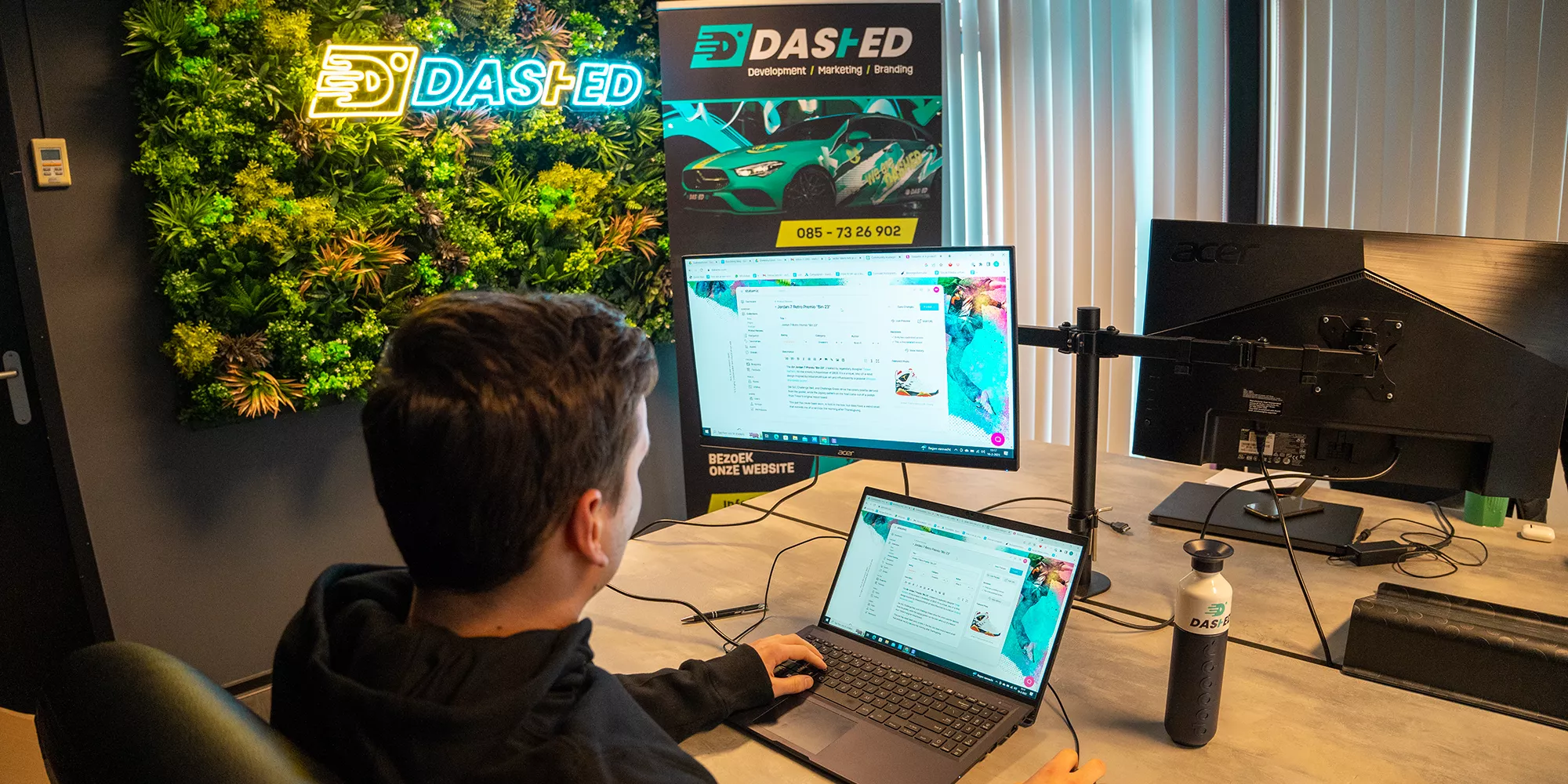Een persoon werkt op een laptop en een groot scherm in een modern kantoor met een groene wand en een poster van een auto met "Dashed" branding.