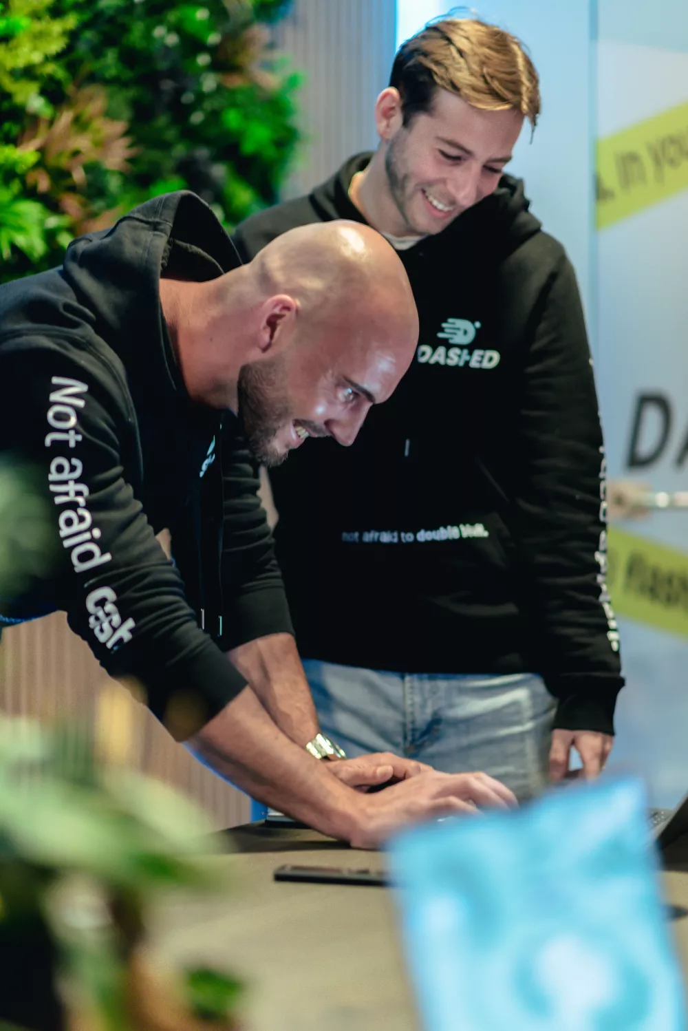 Twee mannen in zwarte hoodies lachen en werken samen aan een laptop bij een bureau met groene planten op de achtergrond.
