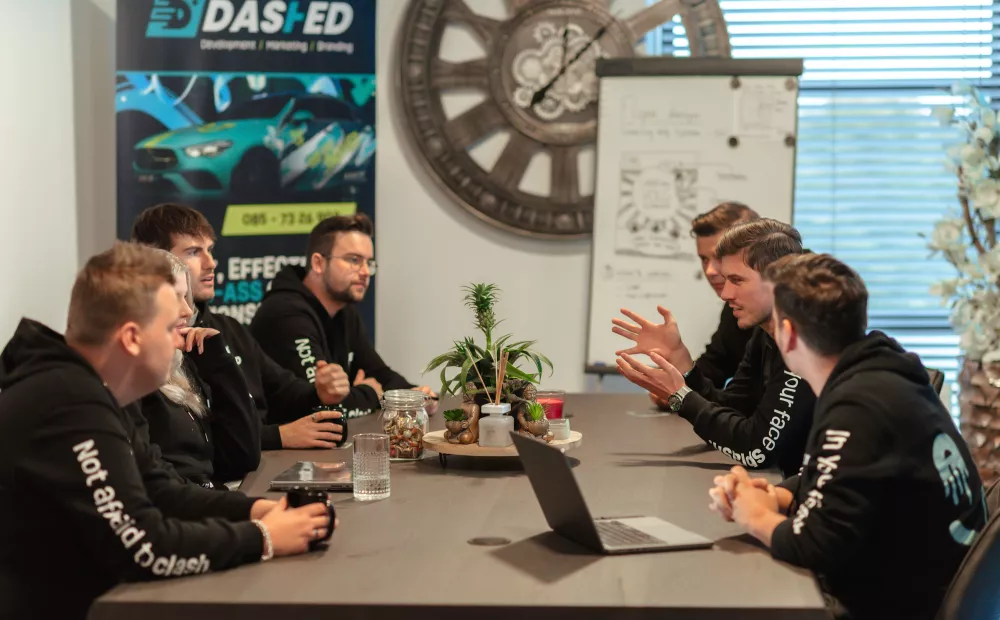Een groep mensen in zwarte hoodies zit aan een vergadertafel in discussie, met een laptop en planten in het midden. Op de achtergrond is een flip-over en een grote klok te zien.
