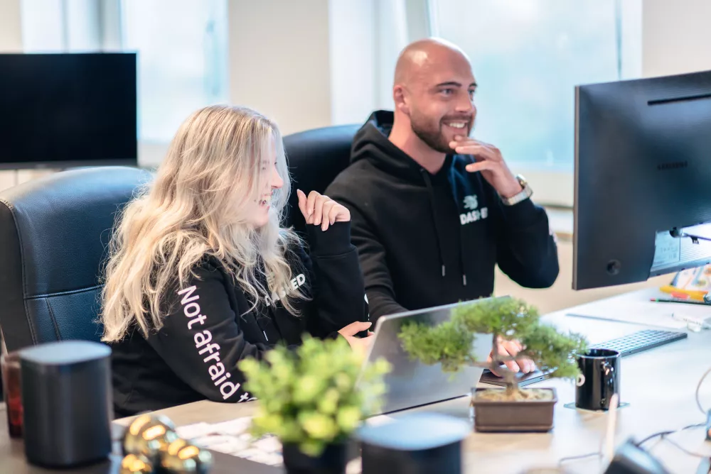 Twee mensen lachen en kijken naar een computerscherm in een kantooromgeving, met planten op het bureau.
