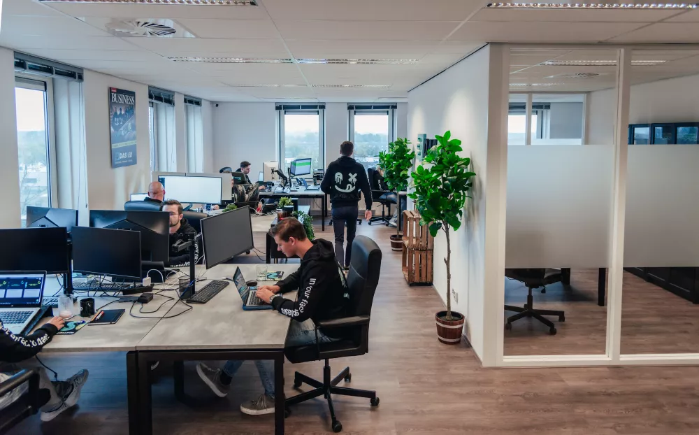 Een modern kantoor met meerdere mensen die aan bureaus werken met computers. Planten en grote ramen zijn zichtbaar.