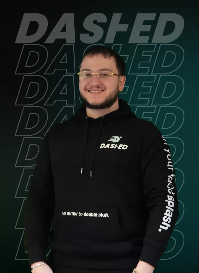 Man met baard en bril draagt een zwarte hoodie met de tekst "DASHED." Achtergrond heeft herhaalde tekst "DASHED."
