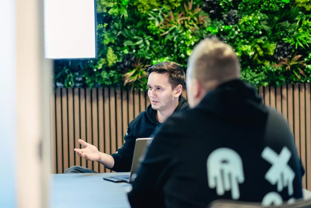 Twee mensen in een vergadering, zittend aan een tafel met laptops. De achtergrond is een groene plantenwand.